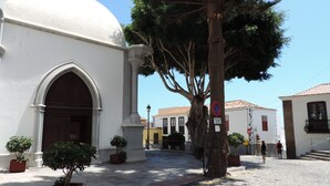 A street with a white building featuring a large arched doorway and a dome. There are potted plants and a tree in the foreground. - Casa Los Helechos (Agulo)
