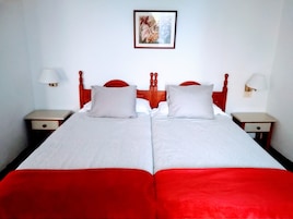 Una cama doble con colcha roja y blanca, dos almohadas, cabecera de madera, dos mesitas de noche con lámparas, y un cuadro en la pared.