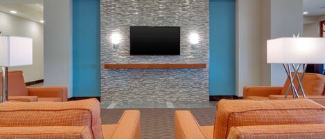 Un vestíbulo de hotel con pared de piedra, un televisor de pantalla plana, asientos tapizados en naranja y una lámpara.