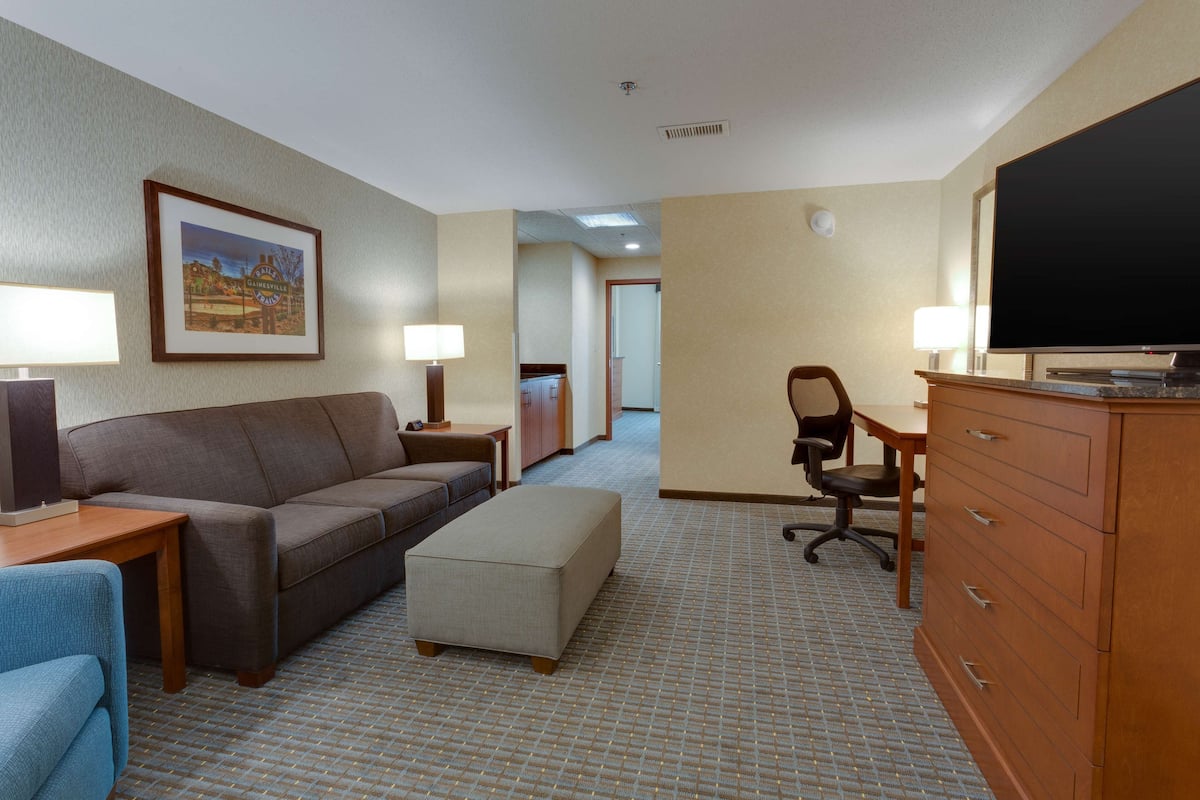A hotel room with a sofa, ottoman, desk, chair, TV, dresser, and framed picture on the wall.