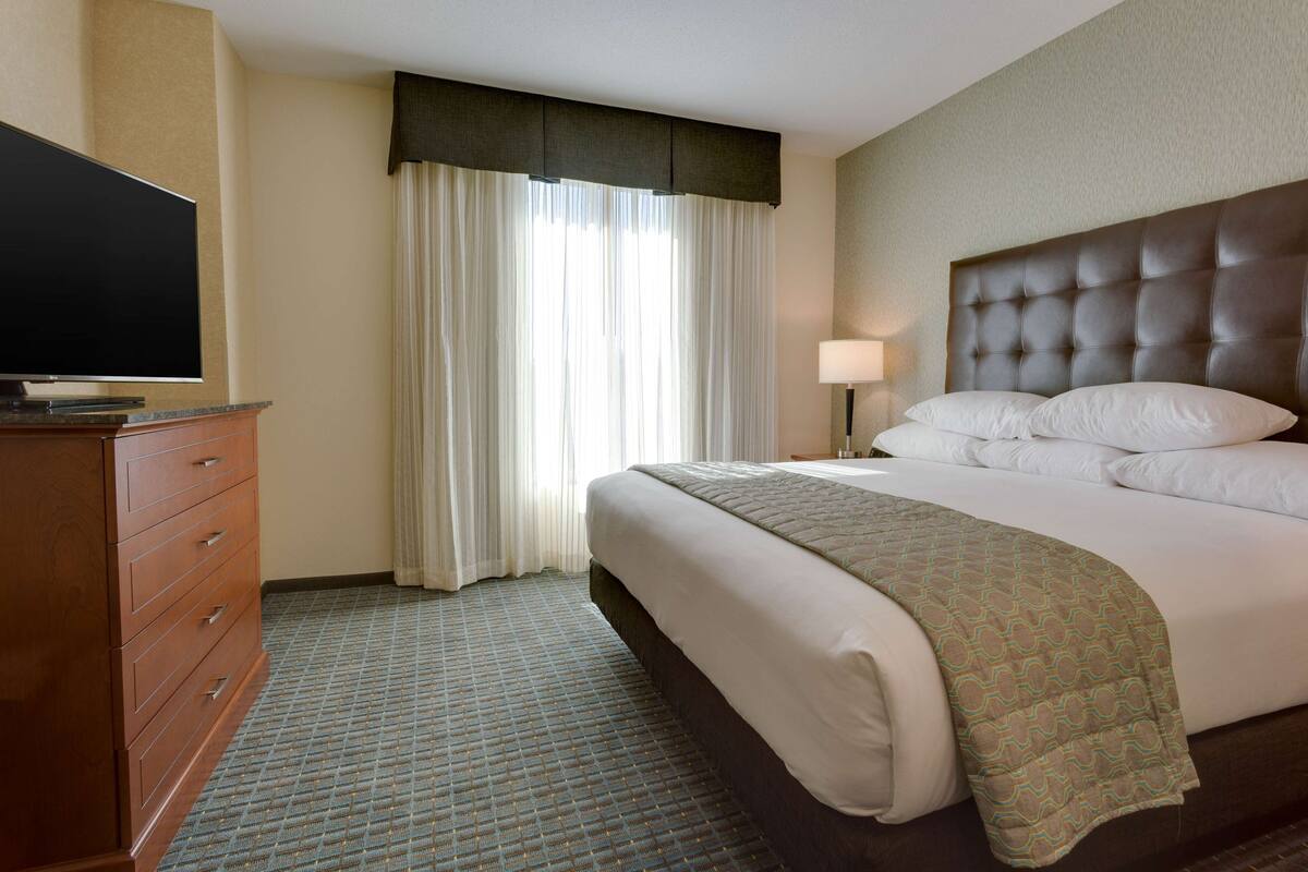 A hotel room with a bed, a television on a wooden dresser, a window with curtains, and a bedside lamp.