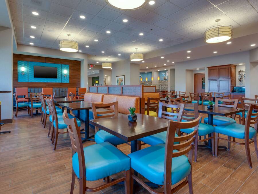 A spacious dining area with multiple tables and chairs, a flat-screen TV mounted on the wall, and a kitchen area in the background.