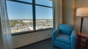 A hotel room with a large window offering a view of a cityscape, a blue armchair, and a small table with a lamp. - Drury Inn & Suites Gainesville (Gainesville)