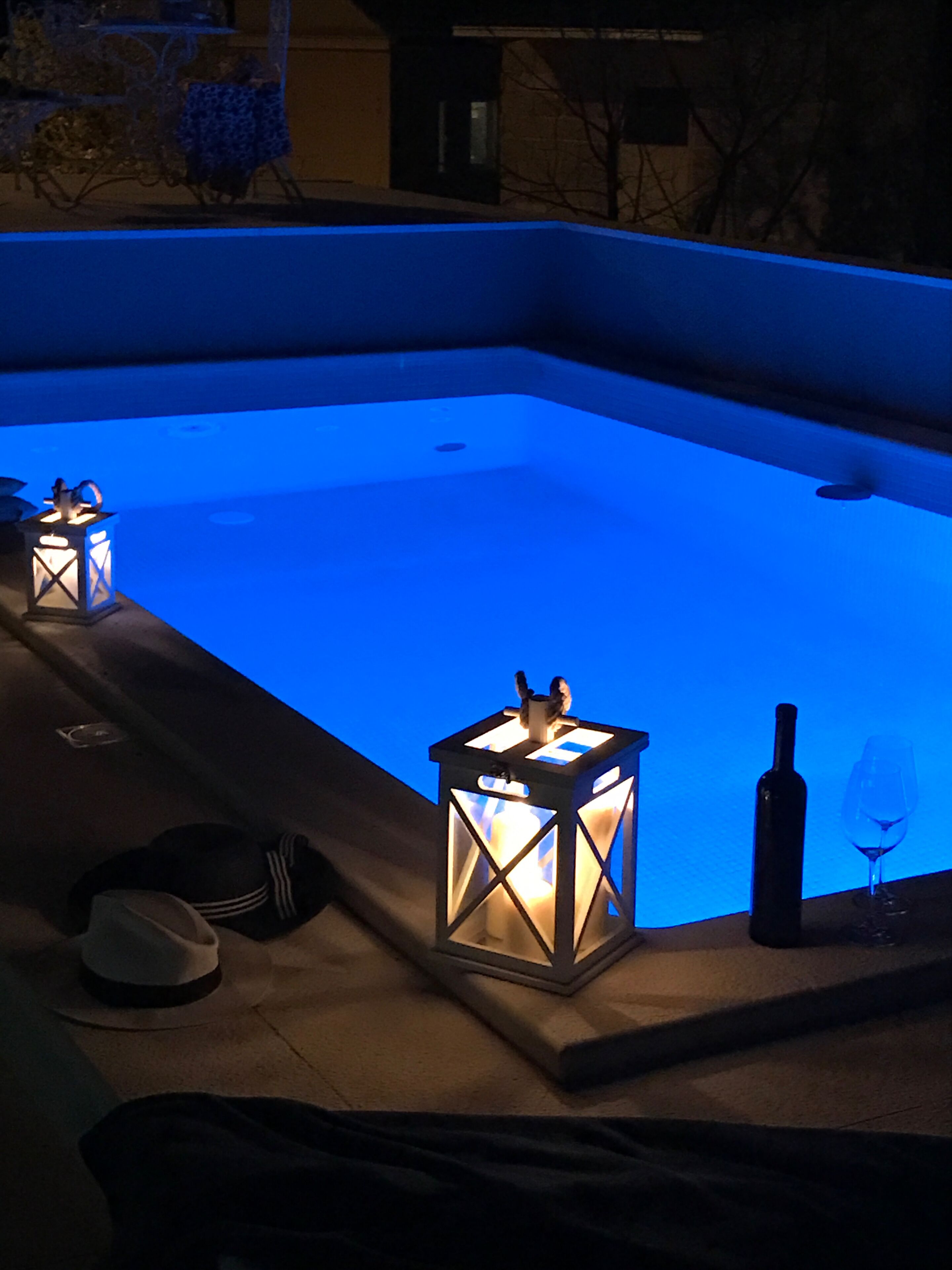 Ein Swimmingpool mit blauer Beleuchtung, umgeben von einer Liegefläche mit zwei Laternen und einer Weinflasche.