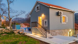 Une maison à deux étages, avec un toit recouvert de tuiles rouges, des fondations en pierre et un petit porche à l’avant, doté d’une balustrade noire.