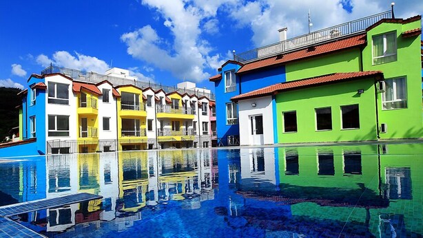 A swimming pool reflecting colorful buildings with balconies, under a clear blue sky. - Large Deluxe Studio in MiniSmart RedZebra Complex (Balchik)