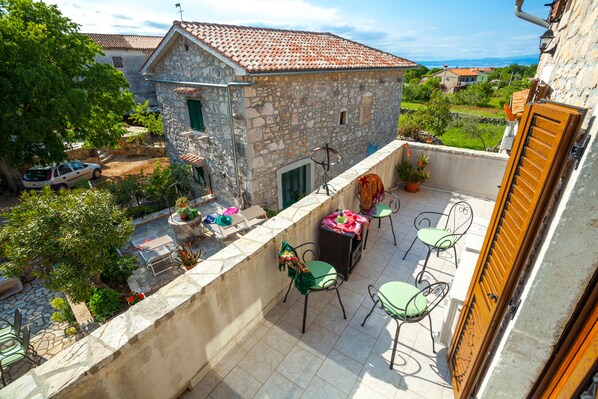 A stone building with a tiled roof, a balcony with green chairs and a table, and a view of the surrounding area.