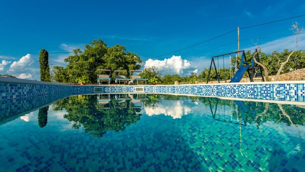 Une piscine d’un bleu éclatant, bordée de carreaux, entourée de verdure et d’un mur de pierre.