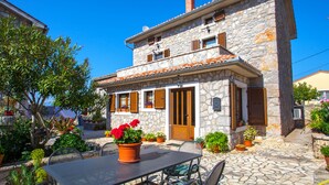 Une maison en pierre avec un toit en tuiles, des volets en bois et un petit porche doté de sièges pour l’extérieur.