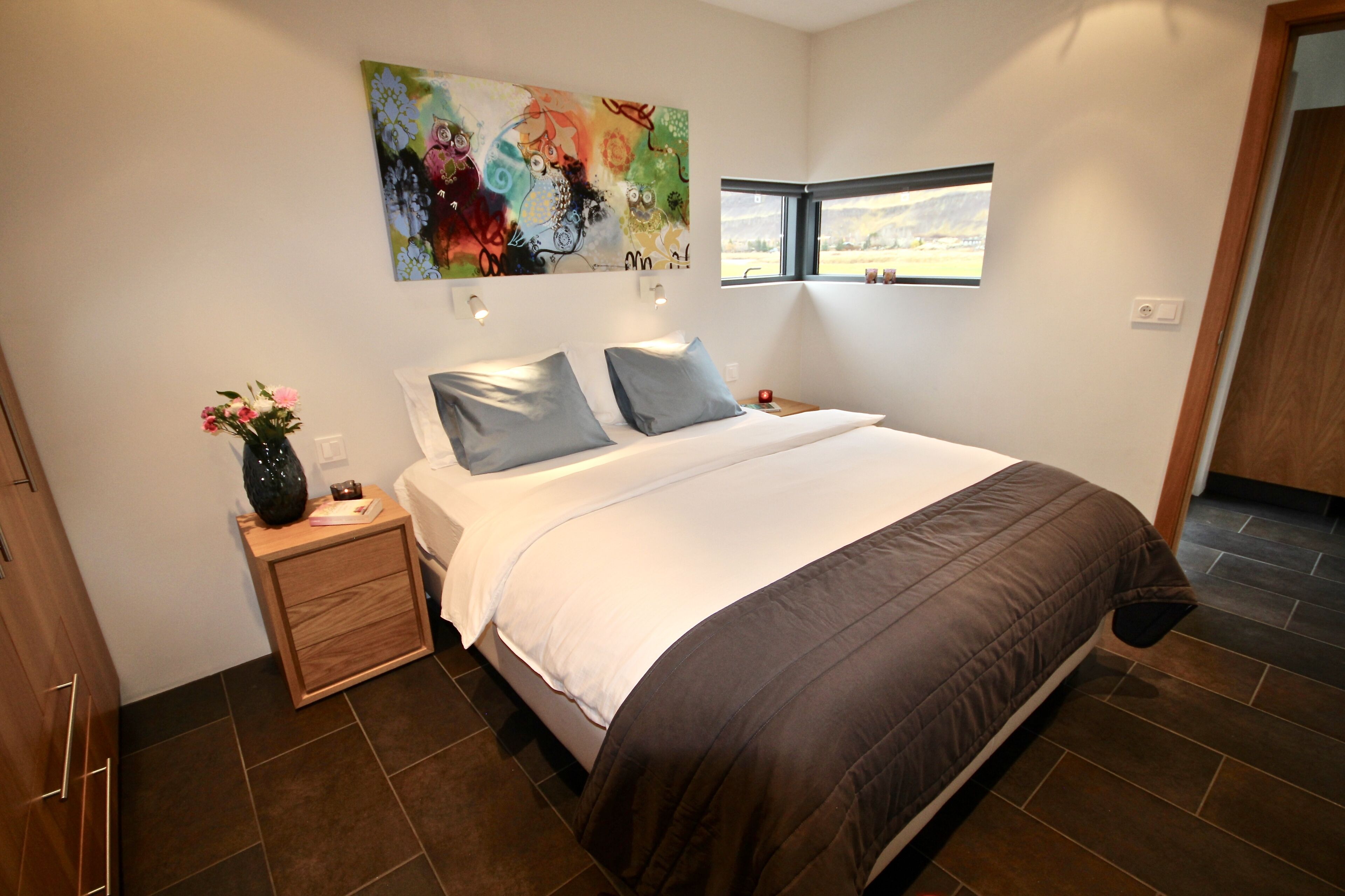 A bedroom with a large bed, two bedside tables, a nightstand, a painting, and a window.