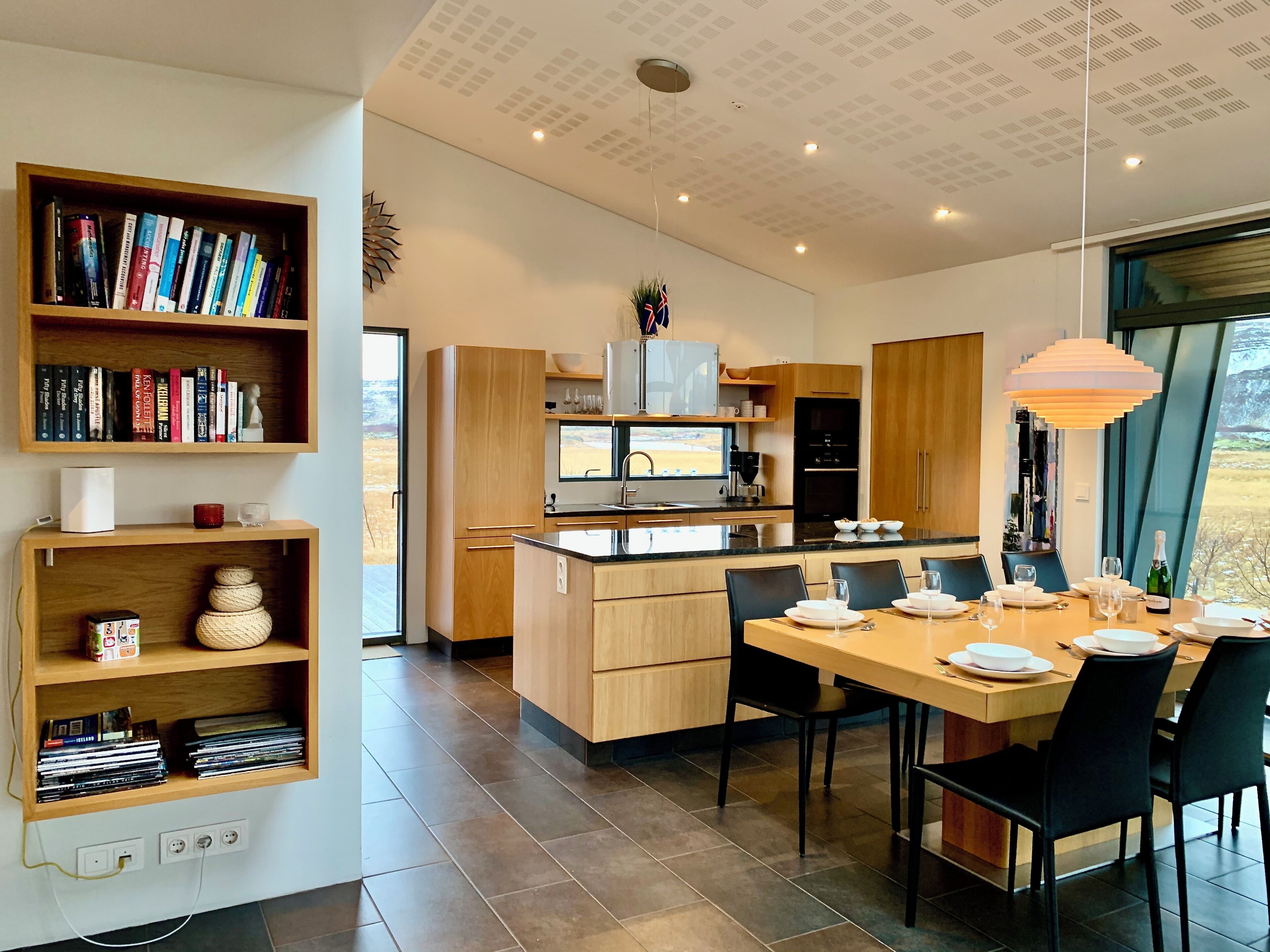 Una cocina moderna con armarios de madera, un comedor con una mesa preparada para cuatro personas, y una estantería llena de libros.