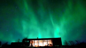 A building with large windows, a visible interior, and a snowy landscape under a night sky with aurora borealis. - Luxury, Modern, Amazing River/Mountain view with Hot Tub. Winter/Summer Holiday (Bifröst)