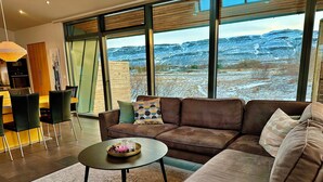 A modern living room with a brown sofa, a coffee table, and a view of a snowy landscape through large windows. - Luxury, Modern, Amazing River/Mountain view with Hot Tub. Winter/Summer Holiday (Bifröst)