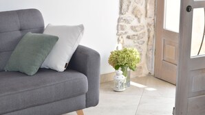 A grey sofa with two pillows, a white vase with green flowers, and a white lantern on a tiled floor near a stone wall and a wooden door. - Charming stone house in Cavtat old town (Cavtat)