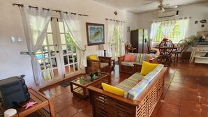 A spacious living area with a wooden sofa, a television, and a dining table. There are curtains on the windows, and a ceiling fan is visible.
