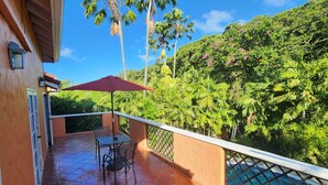 A balcony with a table and chairs, a red umbrella, and a view of lush greenery.