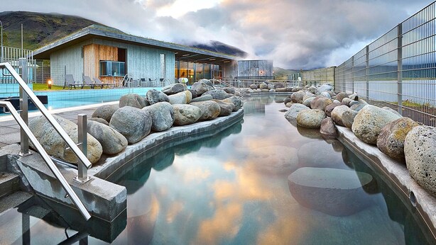 Un espace piscine bordé de grandes roches, un bâtiment à l’extérieur en bois et, au fond, une structure moderne en verre.
