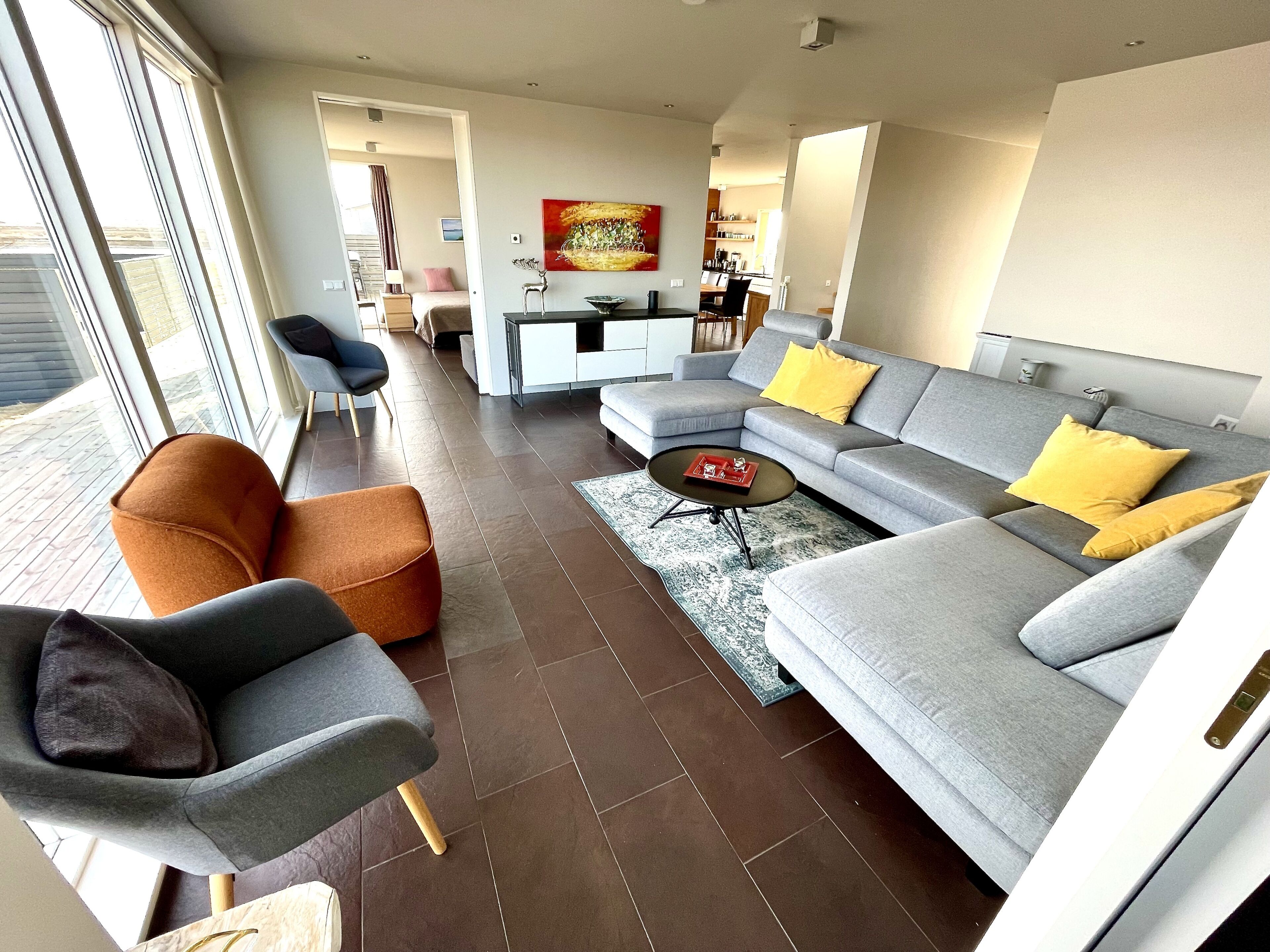 A modern living room with a grey sectional sofa, orange ottoman, and grey armchairs. A coffee table with a book and a rug is in the center. The room has a large window, a kitchen in the background, and a painting on the wall.