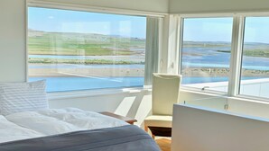 Une chambre à coucher avec un lit, une chaise et une grande fenêtre donnant sur un paysage composé d’étendues d’eau et de collines.