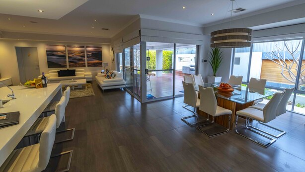 A modern kitchen with a central island, a dining area with white chairs, and a large window offering a view of the outdoors. - Magnificent Beach Retreat (Alkimos)