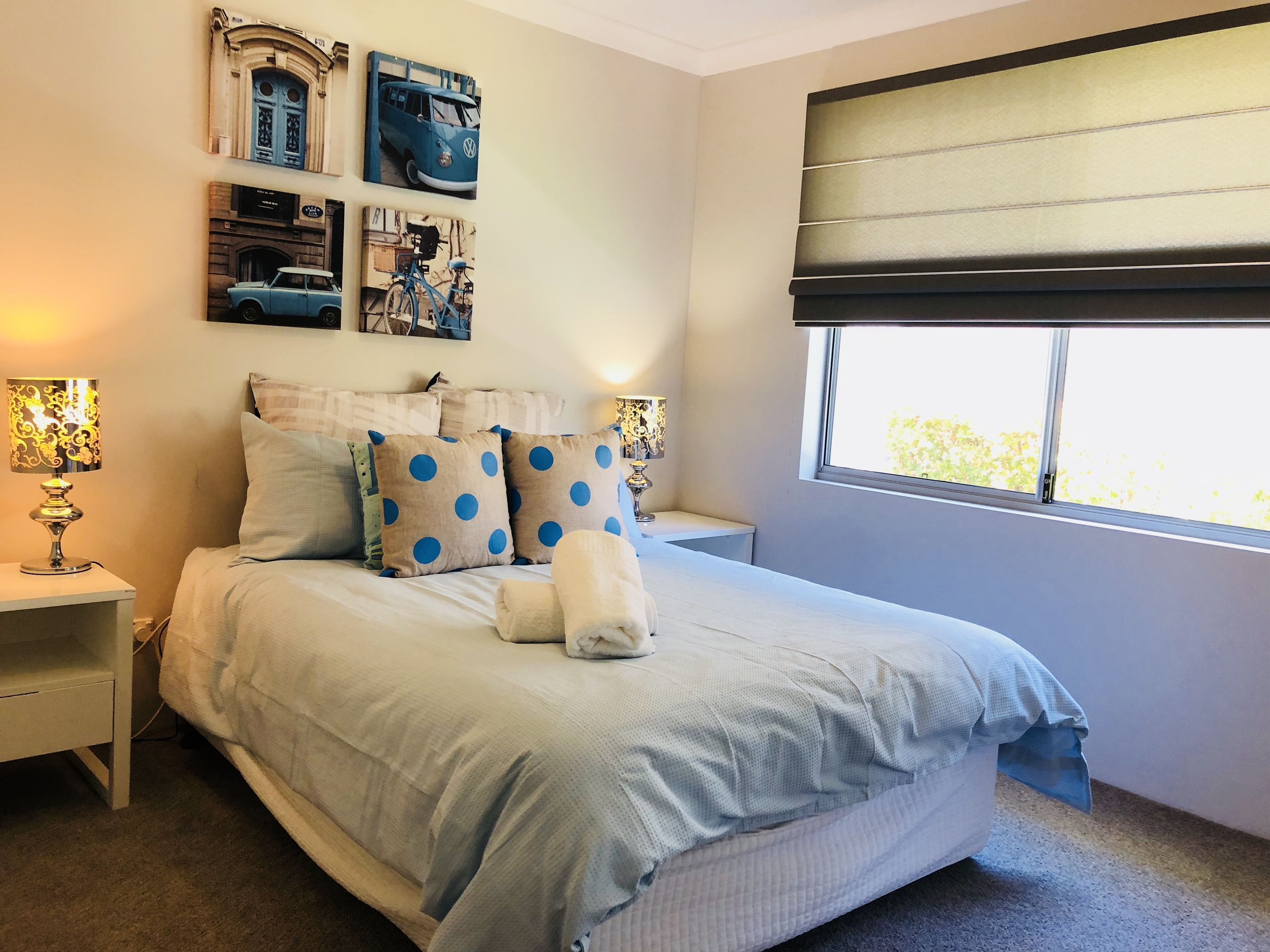 Une chambre à coucher avec un grand lit, deux tables de chevet, une fenêtre avec des stores et des œuvres d’art murales.