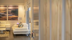 A modern hotel room with a sofa, a small table, and a lamp. There is a large window with blinds, and the room has a neutral color scheme. - Magnificent Beach Retreat (Alkimos)