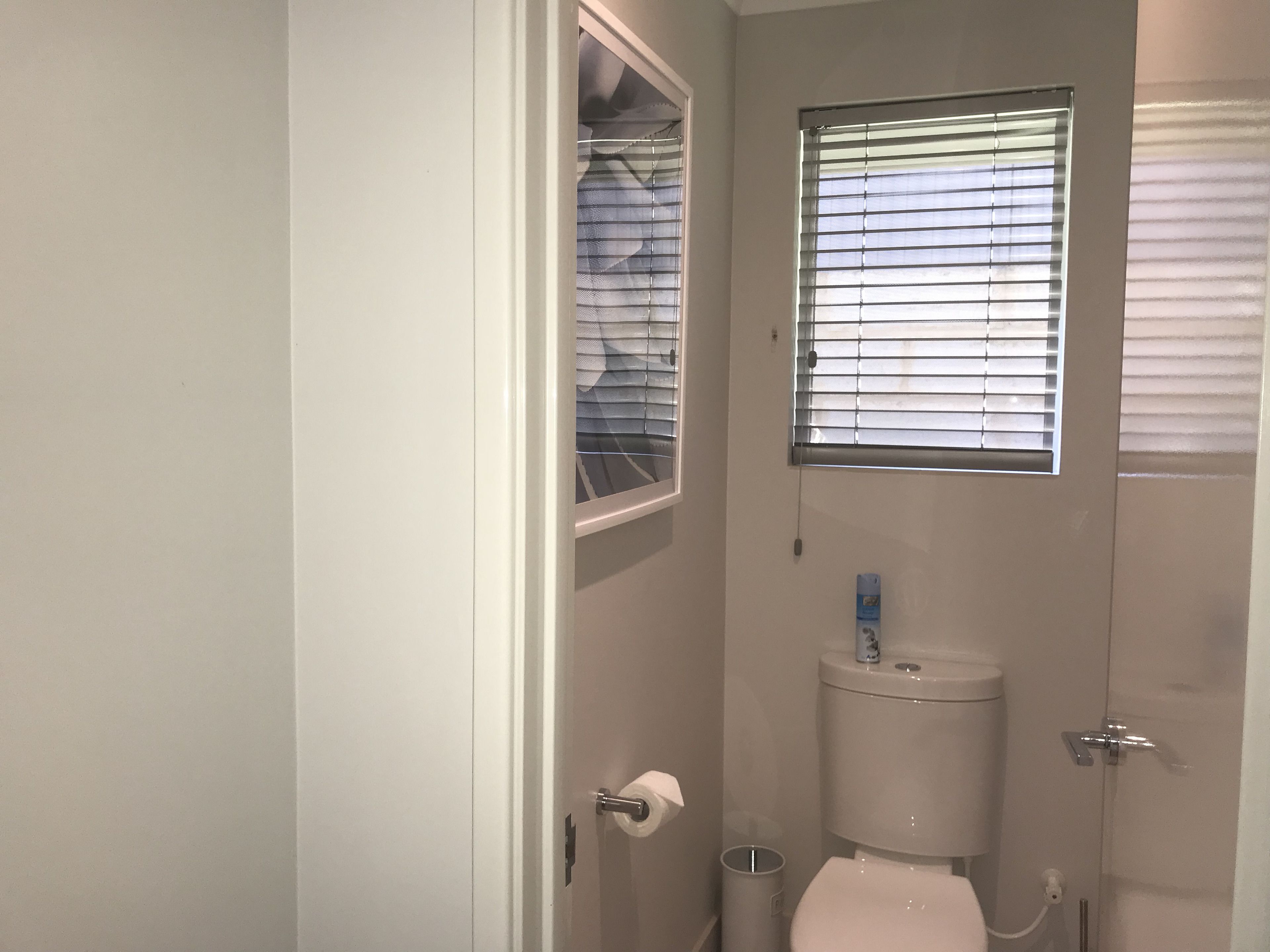 A small, enclosed bathroom with a toilet, a window with blinds, and a shower area.