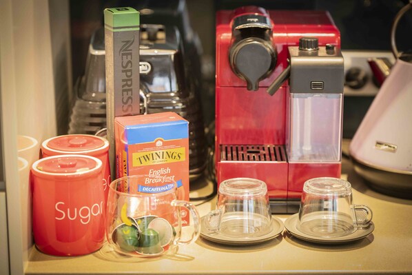 Uma bancada de cozinha com uma cafeteira vermelha, uma máquina Nespresso, recipientes de açúcar, uma caixa de chá Twinings English Breakfast e café descafeinado.