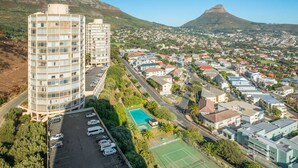 A high-rise hotel with multiple floors, a parking lot, a tennis court, and a view of a town with a prominent mountain in the background. - Iconic Architecture With Sweeping Mountain Views! (Cape Town)