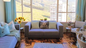 A living room with a sofa, a small table, and a view of a mountainous landscape through large windows. - Iconic Architecture With Sweeping Mountain Views! (Cape Town)
