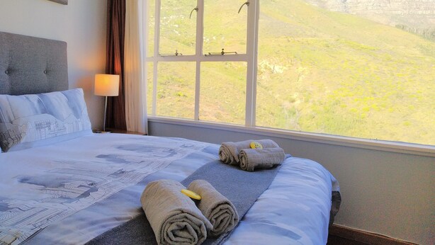A hotel room with a bed, a window with a mountain view, a lamp, and a painting on the wall. - Iconic Architecture With Sweeping Mountain Views! (Cape Town)