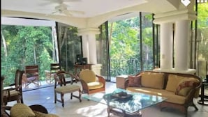 A spacious living area with a glass coffee table, comfortable seating, and large windows offering a view of greenery. - Enchanted Home 7  bedroom home. (Quepos/Manuel Antonio)