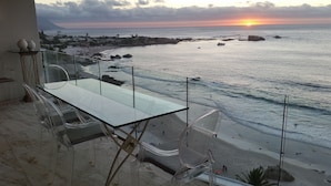 Un balcon avec une table et des chaises en verre, donnant sur une plage et offrant une vue sur le coucher de soleil.
