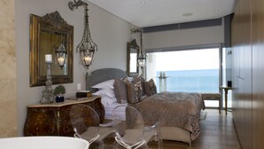 Une chambre avec un grand lit, deux chaises transparentes, une coiffeuse avec un miroir et une vue sur la mer.