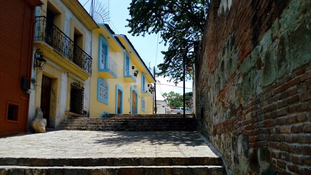 A street with stone steps leading to a yellow building with blue windows and a brick wall on the right. - Casa La Joya in Down Town- Santo Domingo Church (Oaxaca)
