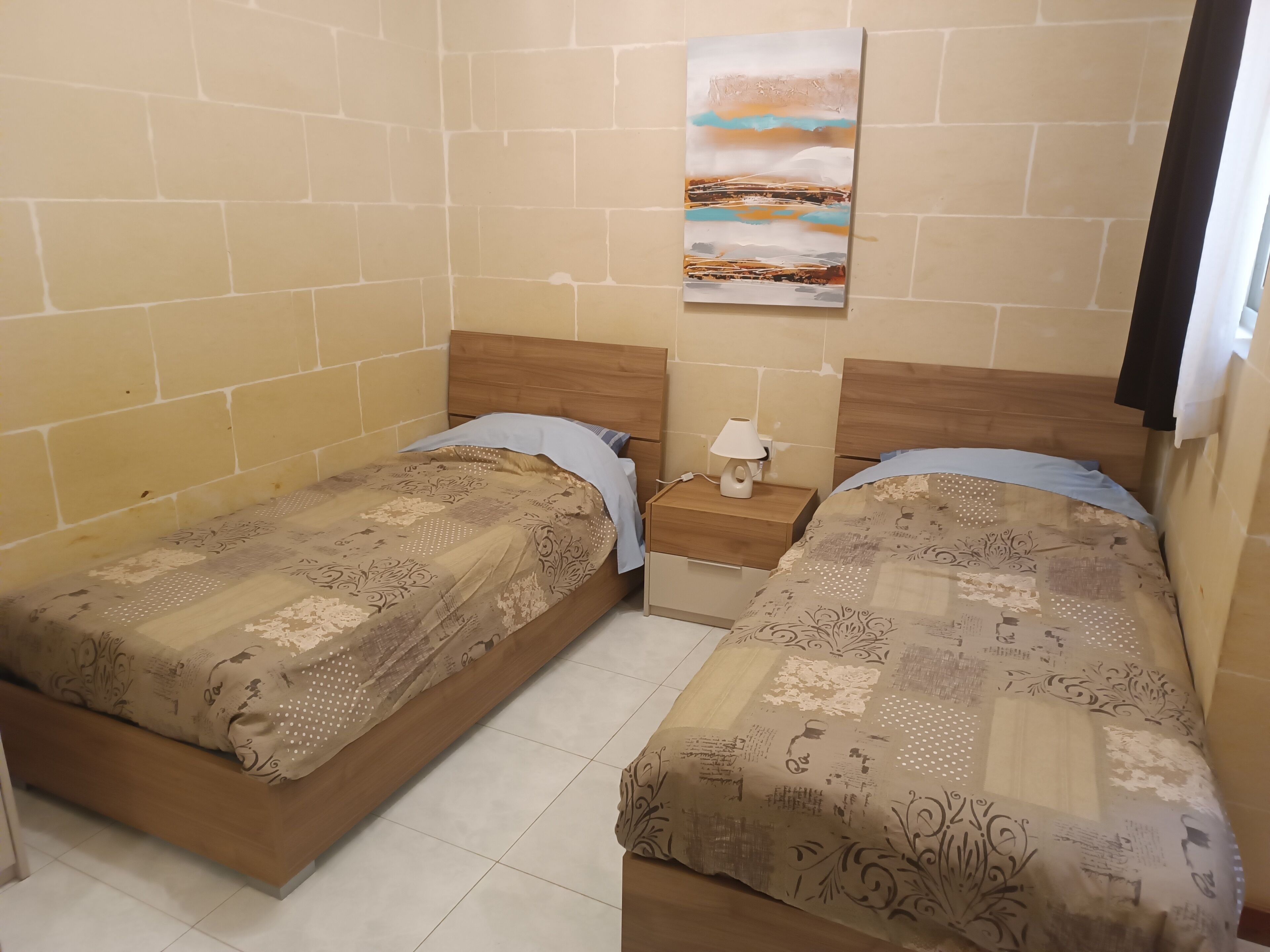 A room with two beds, each with patterned bedding and wooden headboards. There is a nightstand with a lamp between the beds.