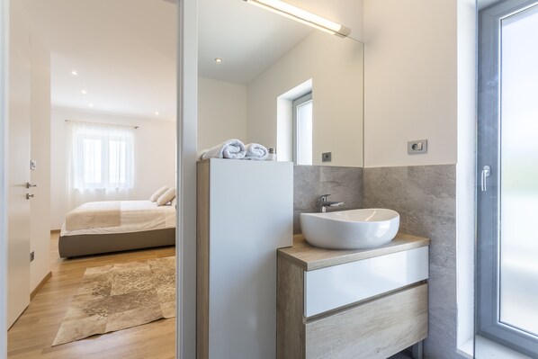 A modern bedroom with a large bed, a bedside table with towels, and a bathroom with a white sink and mirror.