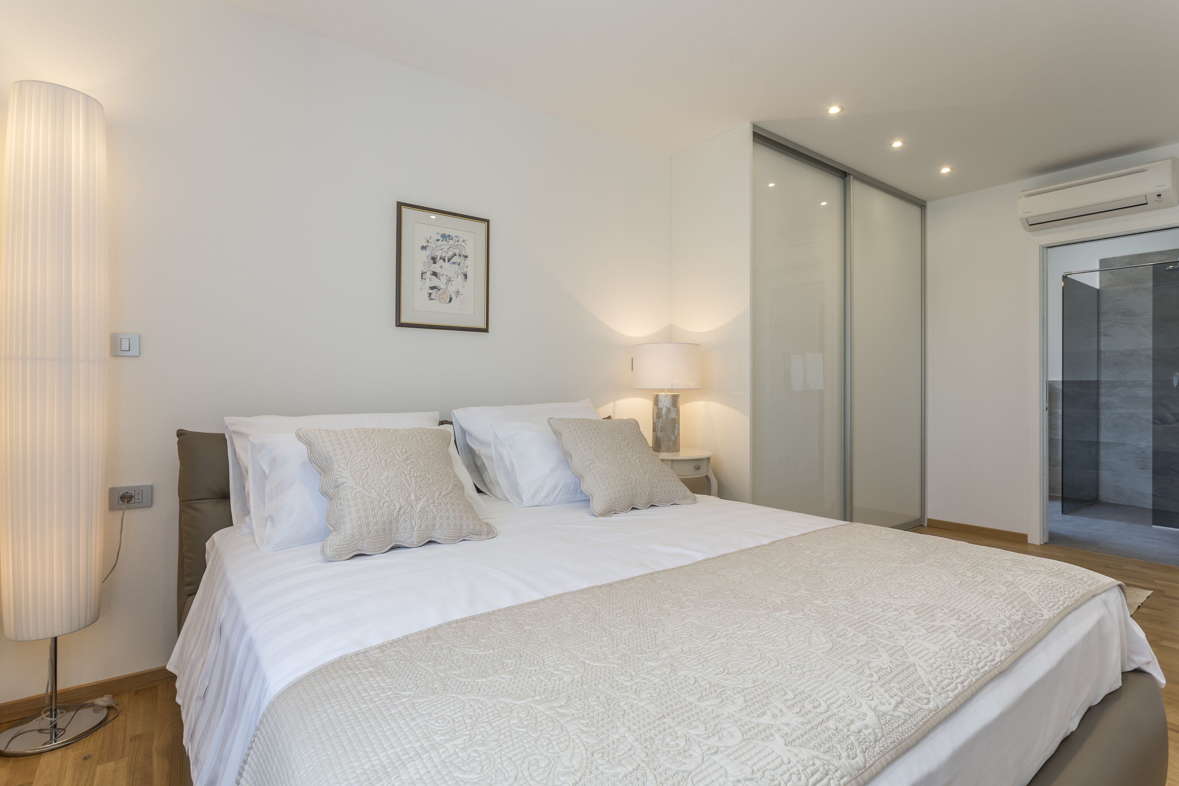 Un dormitorio moderno con una cama grande, una lámpara de noche, un cuadro enmarcado en la pared y una puerta corredera a un balcón.