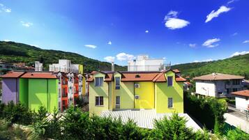 A row of colorful buildings with flat roofs, surrounded by greenery and hills in the background.