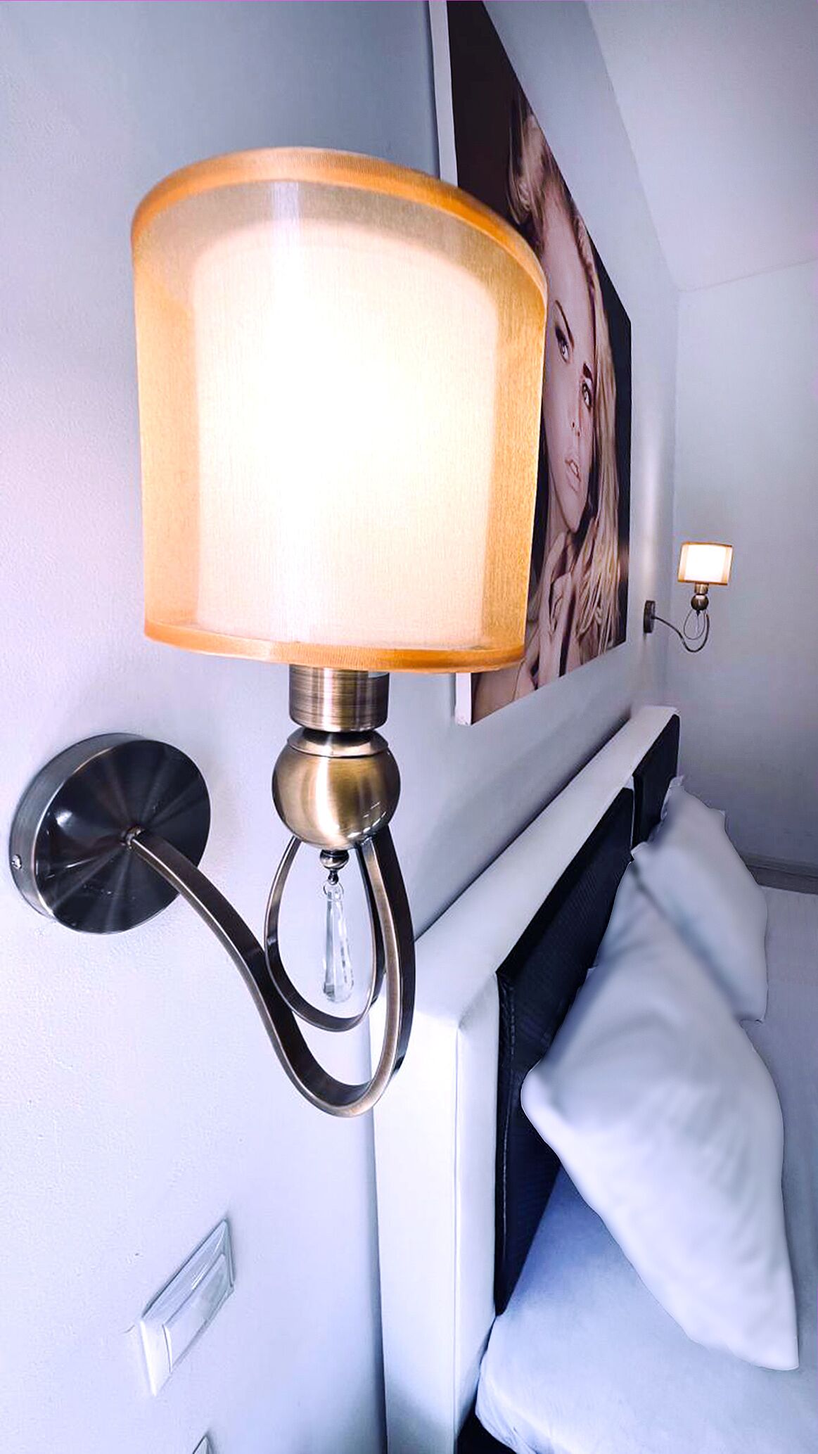 A wall-mounted lamp with a frosted glass shade, a round wall sconce, and a framed picture above a bed with white pillows.