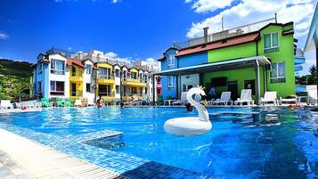 Une piscine avec un flotteur en forme de cygne, entourée de bâtiments aux couleurs vives et de chaises longues.