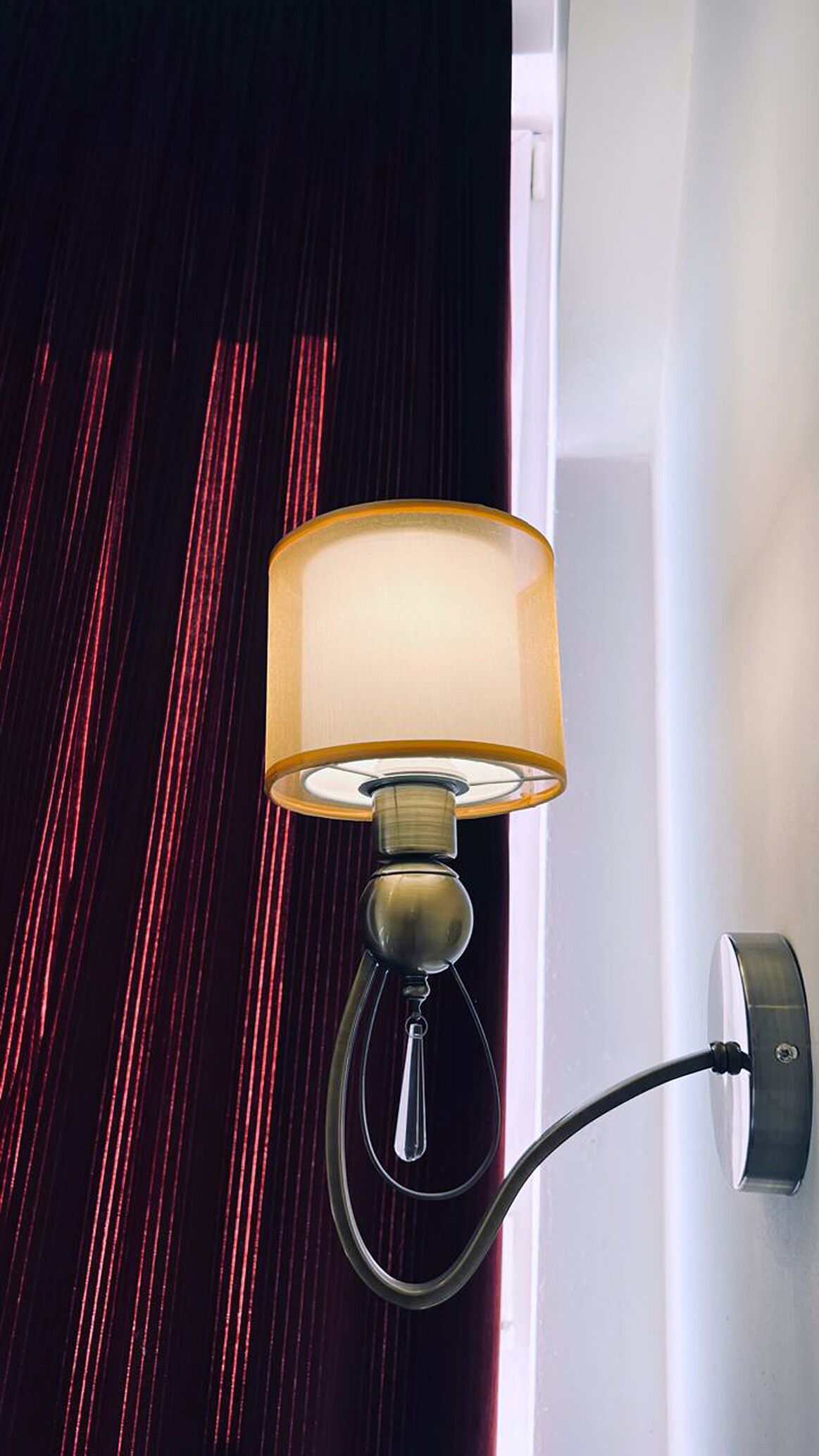 Een wandlamp met een cilindrische lampenkap en een verstelbare arm, bevestigd aan een muur met een rood gordijn op de achtergrond.