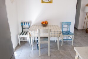 A dining area with a checkered tablecloth and two chairs, one with a blue seat and the other with a white seat, features a basket of oranges on the table.