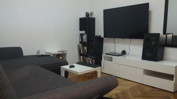 A modern living room with a grey sofa, a white coffee table, a flat-screen TV mounted on the wall, and a bookshelf with books and decorative items.