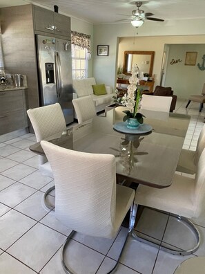 Un espace repas moderne avec une table en verre, des chaises blanches rembourrées, un ventilateur de plafond et une cuisine équipée d’un réfrigérateur en acier inoxydable.