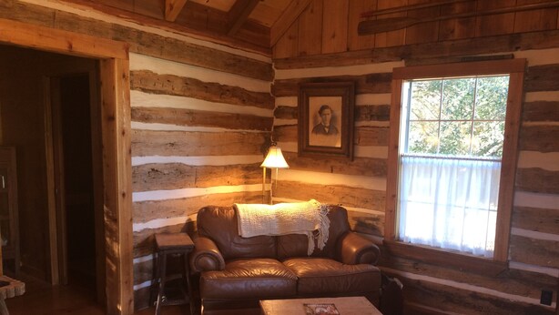 A log cabin interior with a leather sofa, a lamp, a framed portrait, and a window with a view of trees. - Secluded Log Cabin for Two in La Grange TX (La Grange)