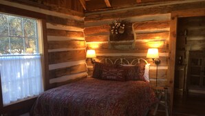 A log cabin bedroom with a bed, two wall-mounted lamps, a decorative wreath, and a window with curtains. - Secluded Log Cabin for Two in La Grange TX (La Grange)