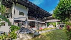 A modern two-story building with a flat roof, large windows, and a covered outdoor area with red chairs. - Luxury Private pool Villa Nap Dau Perfect for Groups (Chalong,Tambon Wichit)