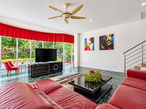 A modern living room with a flat-screen TV, red upholstered sofas, a ceiling fan, and artwork on the walls. - Luxury Private pool Villa Nap Dau Perfect for Groups (Chalong,Tambon Wichit)