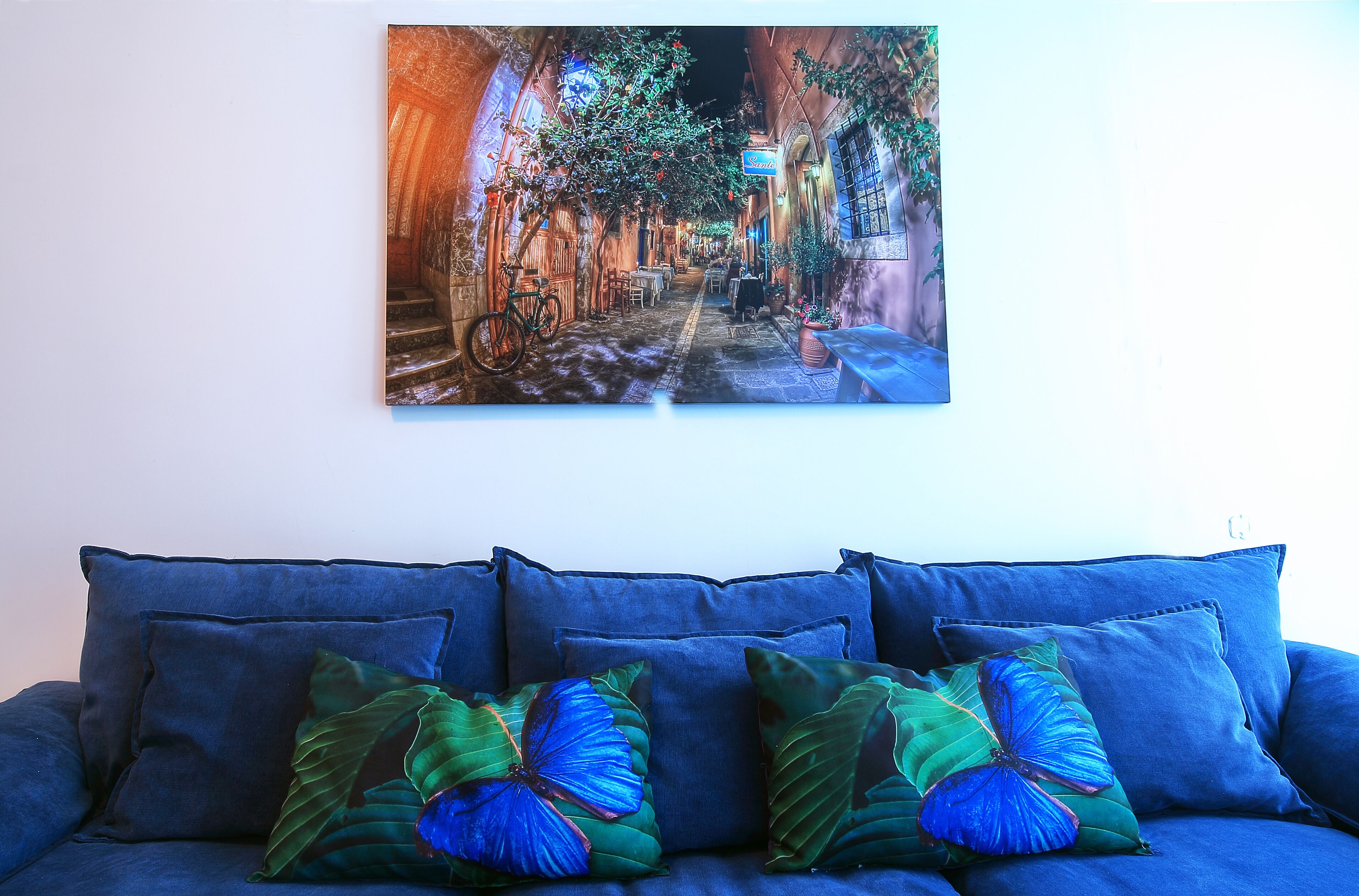 A blue sofa with butterfly-patterned cushions and a painting of a street scene above it.